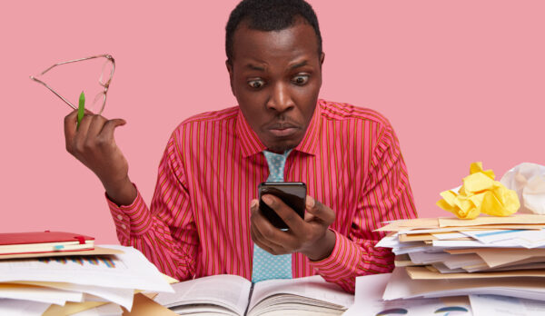 Horizontal shot of emotive stupefied puzzled dark skinned male worker holds mobile phone, stares at screen, recieves message for paying bills, has mess on working table, isolated on pink wall
